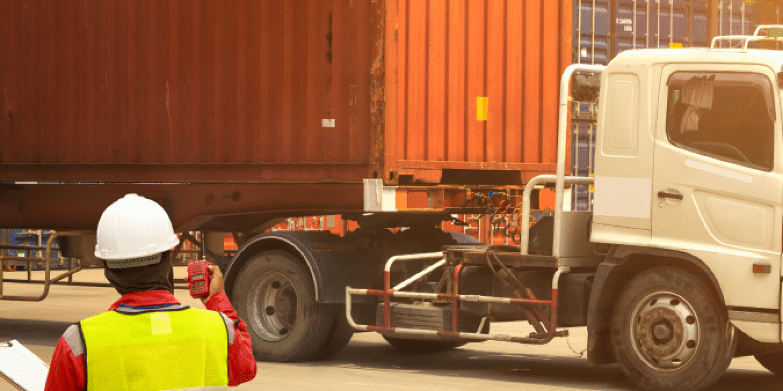 A worker in a reflective vest and hard hat inspects a large shipping container on a truck at a busy shipping yard with stacked containers in the background.