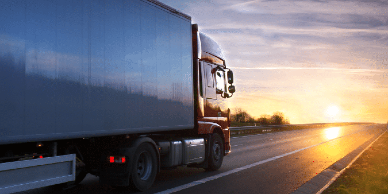 A large semi-truck drives on an empty highway at sunset, with the sky displaying orange and blue hues and the sun low on the horizon.
