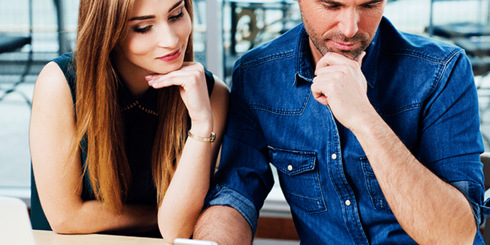 A man and woman sit at a desk, thoughtfully looking at a smartphone and colorful cards arranged on the table. A laptop and pens are nearby, suggesting a collaborative planning or brainstorming session.