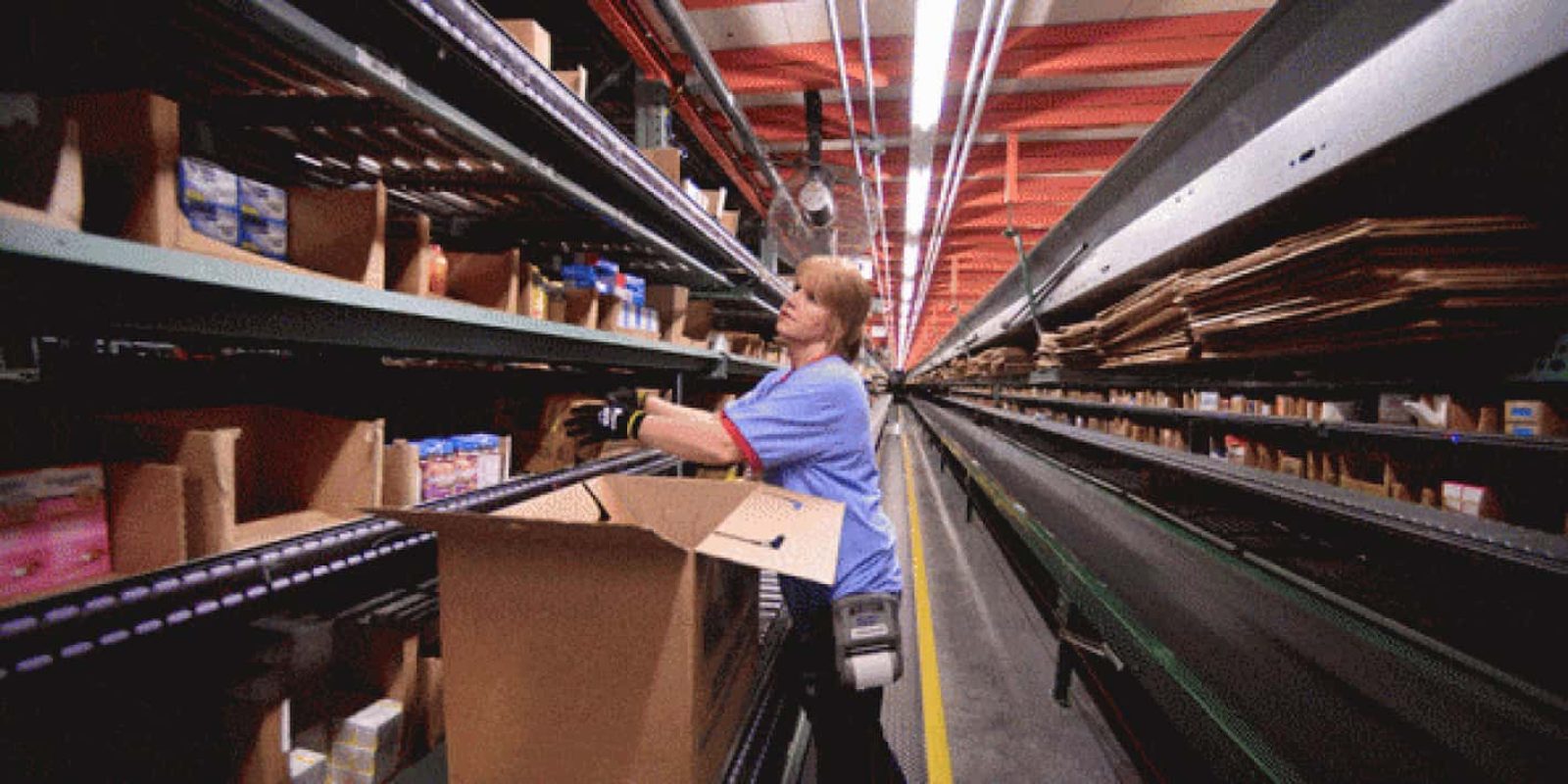 A worker in a blue uniform places products into a large cardboard box on a conveyor belt in a warehouse with shelves filled with various items.