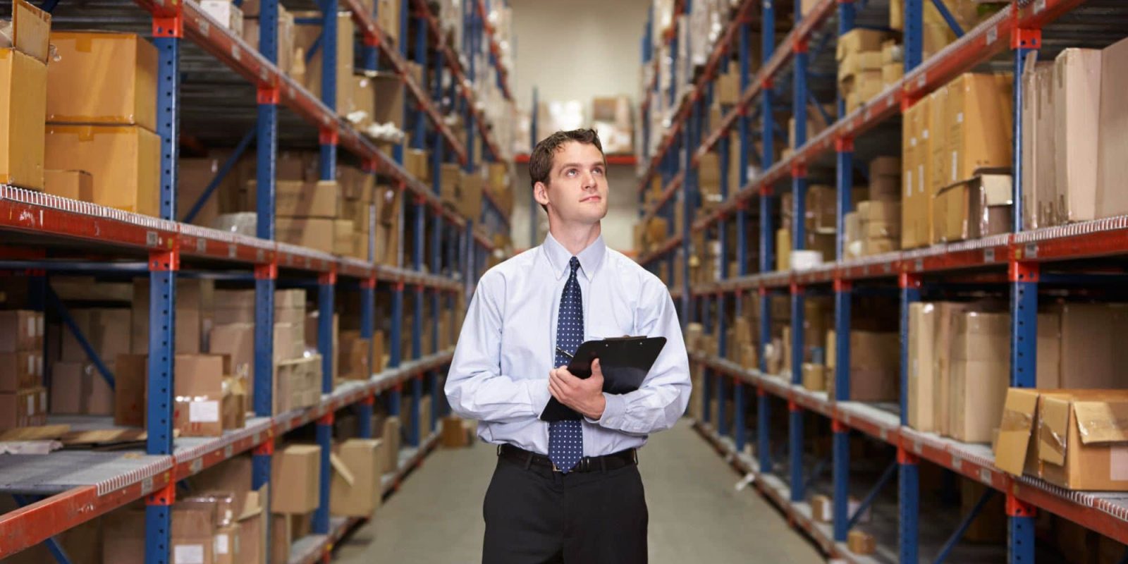 A man in business attire holding a clipboard stands in the aisle of a warehouse, surrounded by tall shelves filled with boxes and packages.