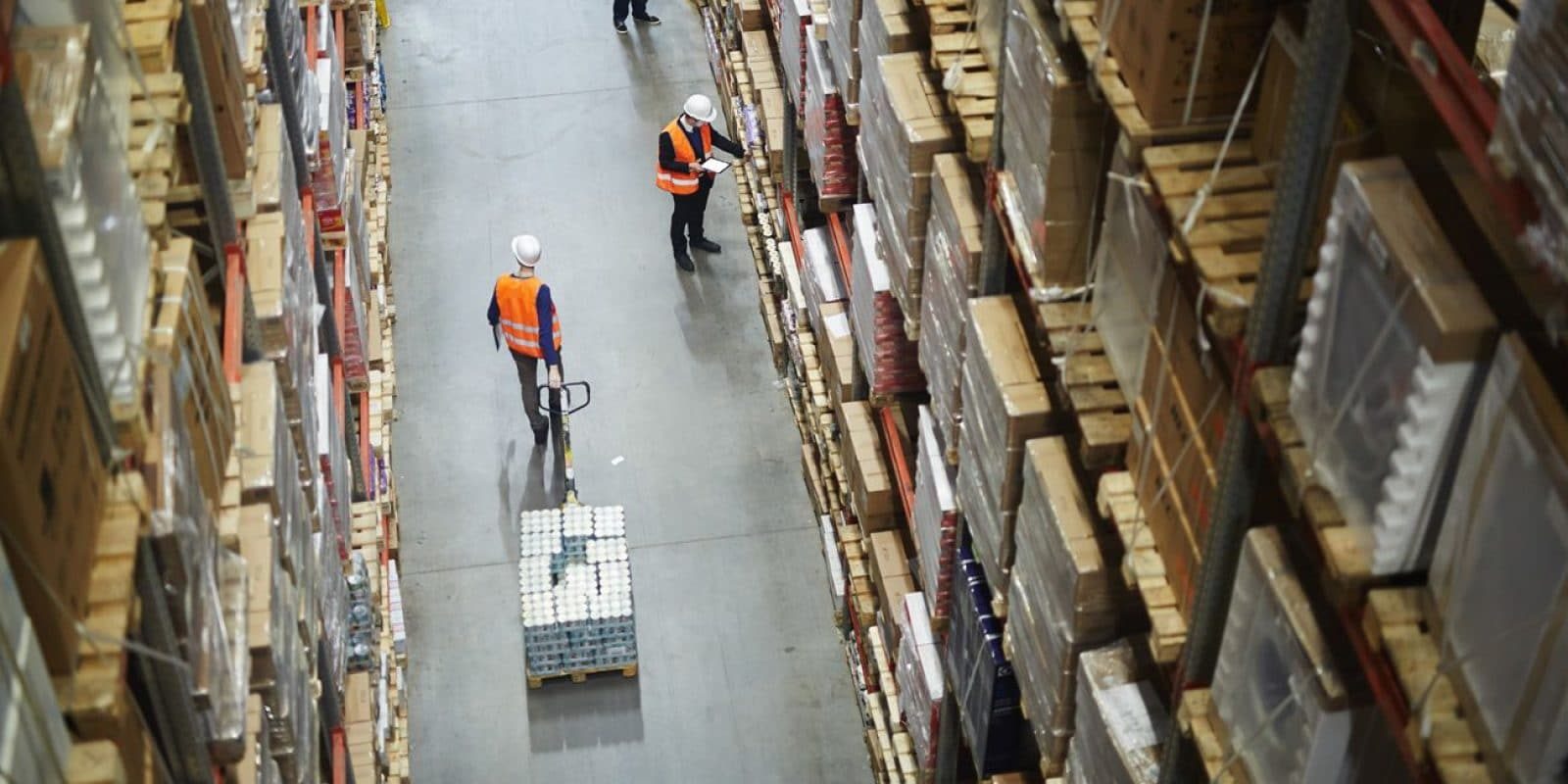 Aerial view of two workers in safety vests and helmets in a warehouse aisle, one pulling a pallet jack with boxes, the other holding a clipboard among tall shelves filled with neatly stacked packages.