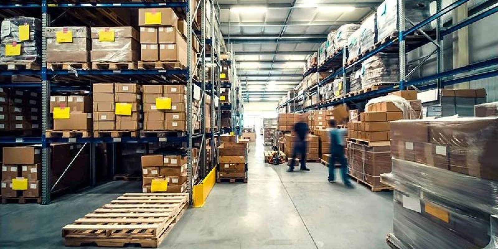 Warehouse interior with tall shelves stacked with cardboard boxes and packages. Two blurred workers, motivated by incentive based pay, are moving around, and pallets are visible in the aisle. The space is brightly lit and organized.